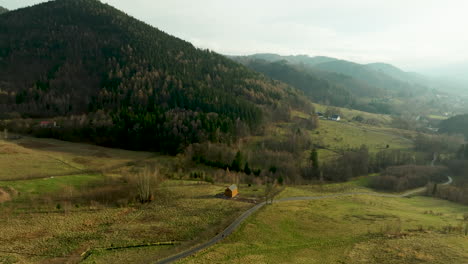 Luftaufnahme---üppiges-Grünes-Tal-Mit-Einer-Kurvenreichen-Straße,-Die-Zu-Einem-Einsamen-Haus-Führt,-Das-Am-Fuße-Einer-Dicht-Bewaldeten-Bergkette-Liegt