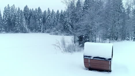 Wooden-Cylindrical-Sauna-Surrounded-by-Falling-Snow-with-Forest-Background,-Aerial-Panning-Drone-Shot