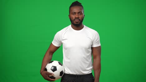 retrato de disparo medio de un hombre afroamericano confiado con una pelota de fútbol posando en el fondo de la pantalla verde. vista frontal de un guapo deportista en una plantilla de chromakey mirando a la cámara. concepto deportivo.