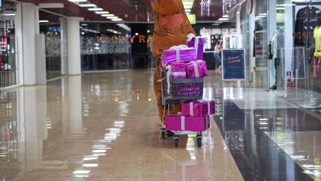 cheerfully running t-rex running by the mall pushing the cart filled with presents. dinosaur getting ready for the holiday in shopping mall.