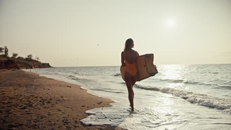 back view: a blonde girl in an orange swimsuit carries a light-colored wooden surfboard and walks along the sandy seashore in the morning at sunrise. a blonde girl surfing master walks along the sea with her surfboard