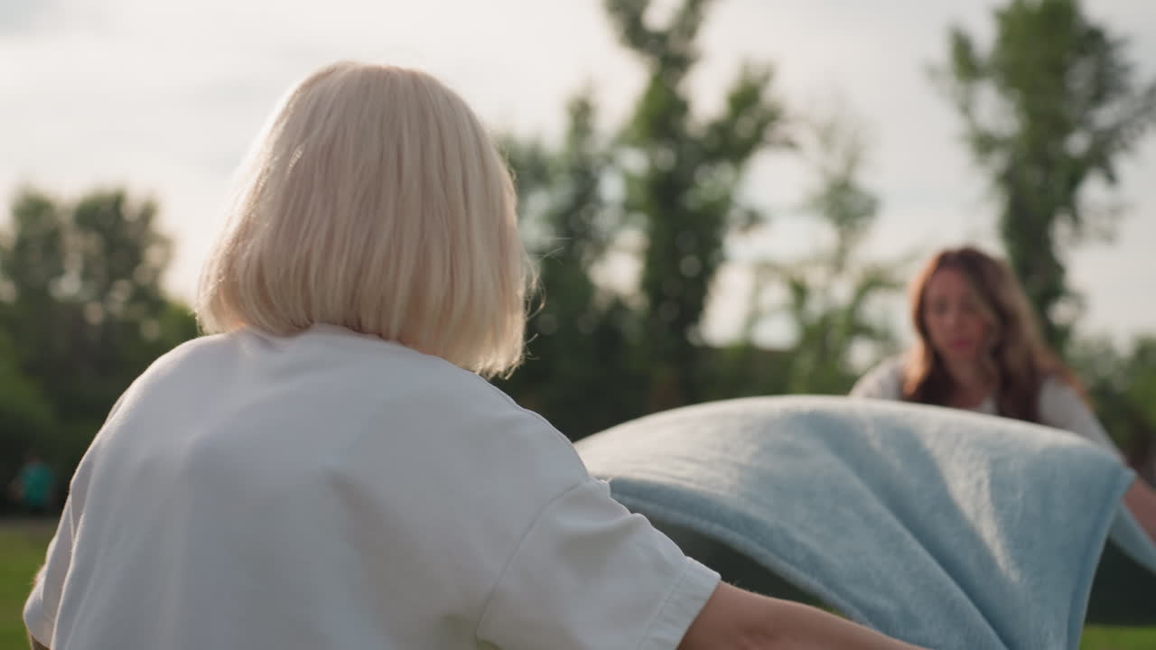 mature women spreading blanket on green grass in park with blurred background, soft sunlight creating warm mood, cooperative gesture preparing area for relaxing gathering