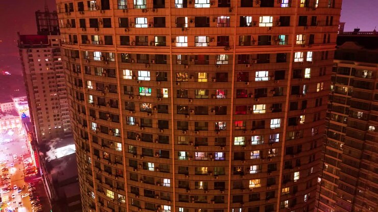 Aerial night view of a modern high-rise apartment building in Harbin, China, with illuminated windows creating a warm, grid-like pattern. The city lights in the background add depth to the urban scene