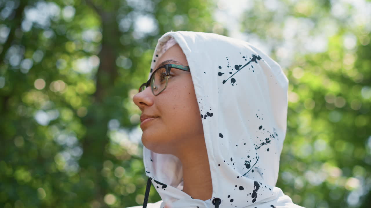 햇빛이 비치는 나무들 사이에서 옆을 바라보는 후드티를 입은 혼혈 여성, 안경을 쓴 측면 샷, 사색에 잠긴 분위기, 후드와 목을 매만지는 손, 평화로운 주변광과 부드러운 숲 배경이 분위기를 자아냅니다.