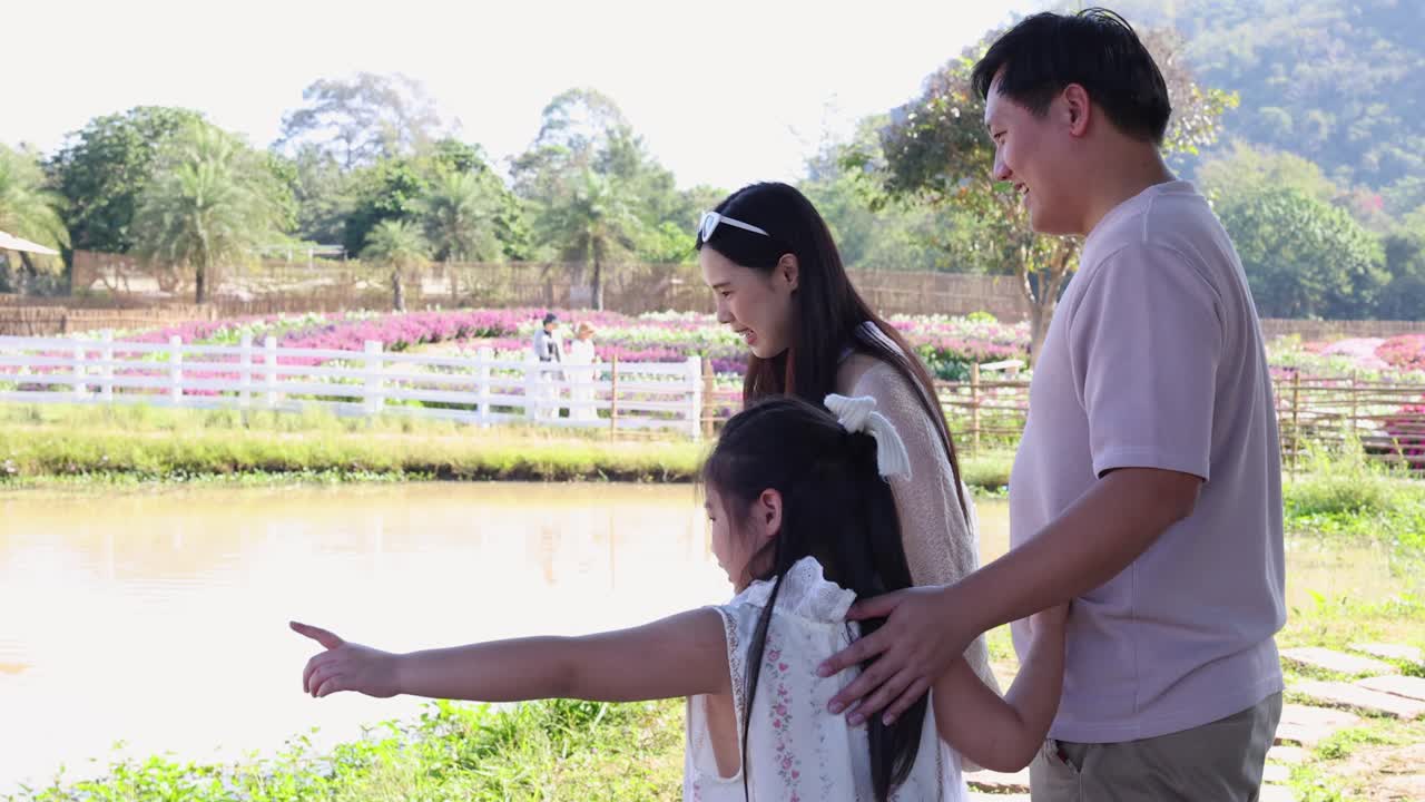 ครอบครัวสุขสันต์เดินเล่นริมทะเลสาบที่มีแสงแดดส่อง เด็กๆ ชี้ไปยังพืชพรรณสีเขียวสวยงามและดอกไม้ที่อยู่ห่างออกไป