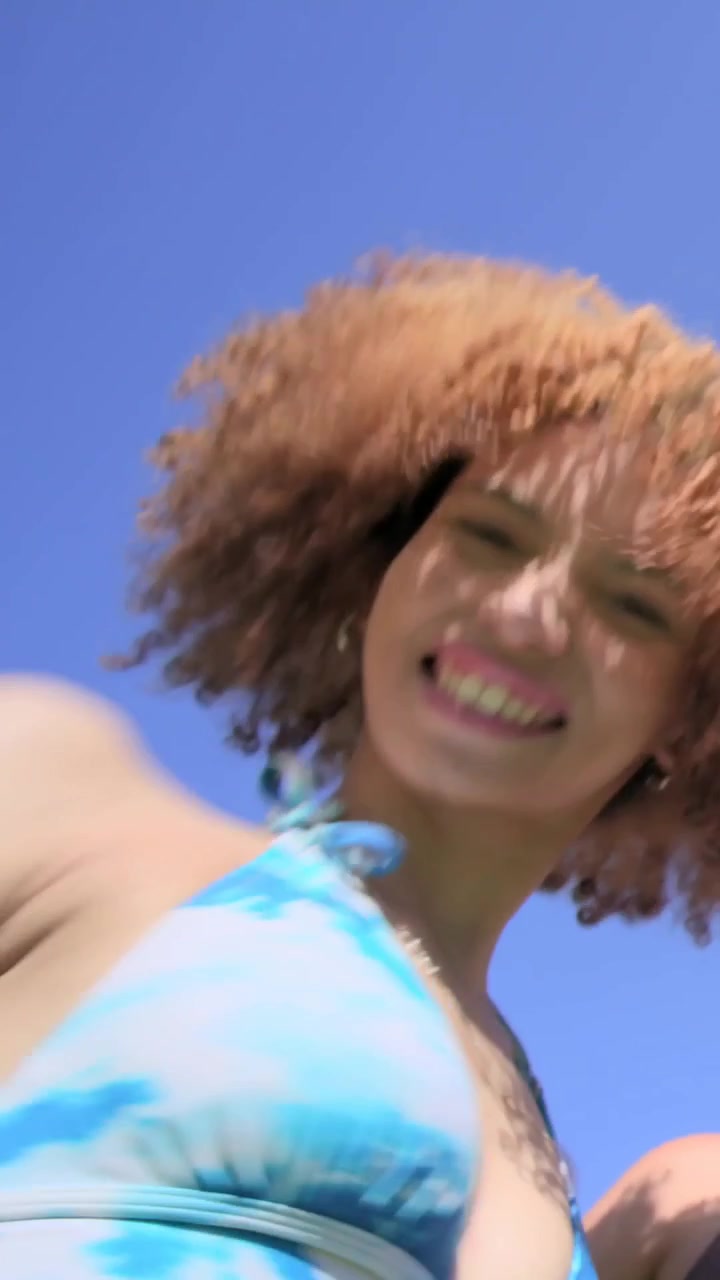 Video verticale: Telefono inclinato che mostra una donna riccia in bikini blu che manda giocosamente un bacio all'obiettivo al parco