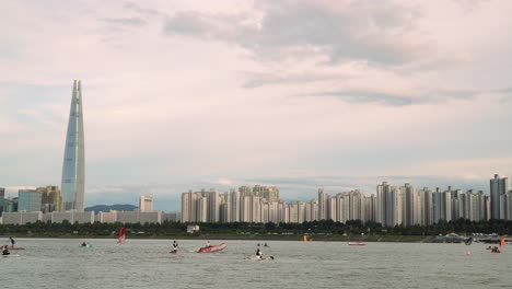 Torre-De-Lotte-En-Corea-Del-Sur---Río-Han-Con-Gente-Disfrutando-Del-Windsurf,-Paddleboarding,-Paseos-En-Bote-Y-Kayak-Con-El-Horizonte-De-Seúl-En-El-Fondo