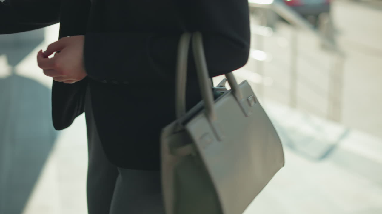 Side view of lady carrying ash handbag walking to office, about to open glass door, dressed in black jacket, professional setting, urban background blurred