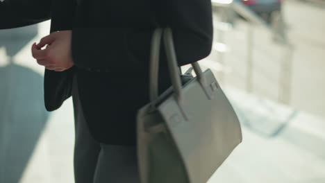 side view of lady carrying ash handbag walking to office, about to open glass door, dressed in black jacket, professional setting, urban background blurred