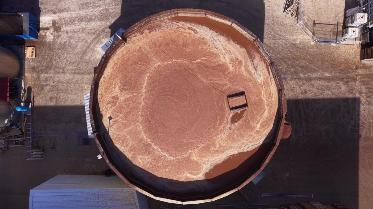 Vista dall'alto di un grande serbatoio chiarificatore industriale circolare riempito con acqua torbida marrone carica di sedimenti, in un impianto di lavorazione circondato da infrastrutture in cemento.