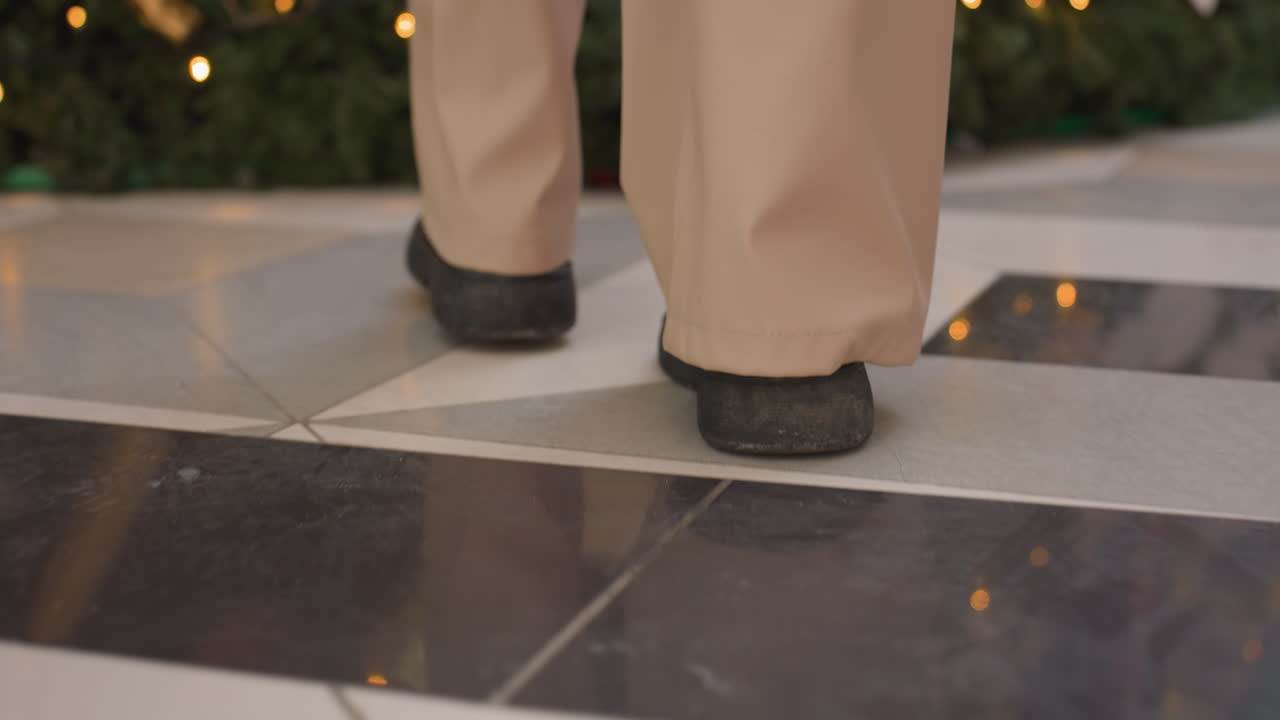 Rear leg view of person wearing black boots walking on well-tied floor towards decorative plant, urban structure in background with light reflections on ground