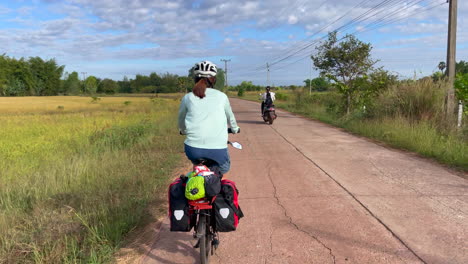 una toma panorámica estacionaria de una mujer que viaja en bicicleta con alforjas pasando por un campo de arroz al borde de la carretera