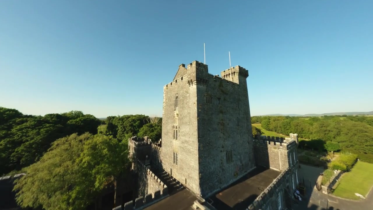 Un volo FPV segue un viale alberato di una tenuta oltre gli alberi prima di elevarsi sopra la torre merlata e i campi verdi circostanti.