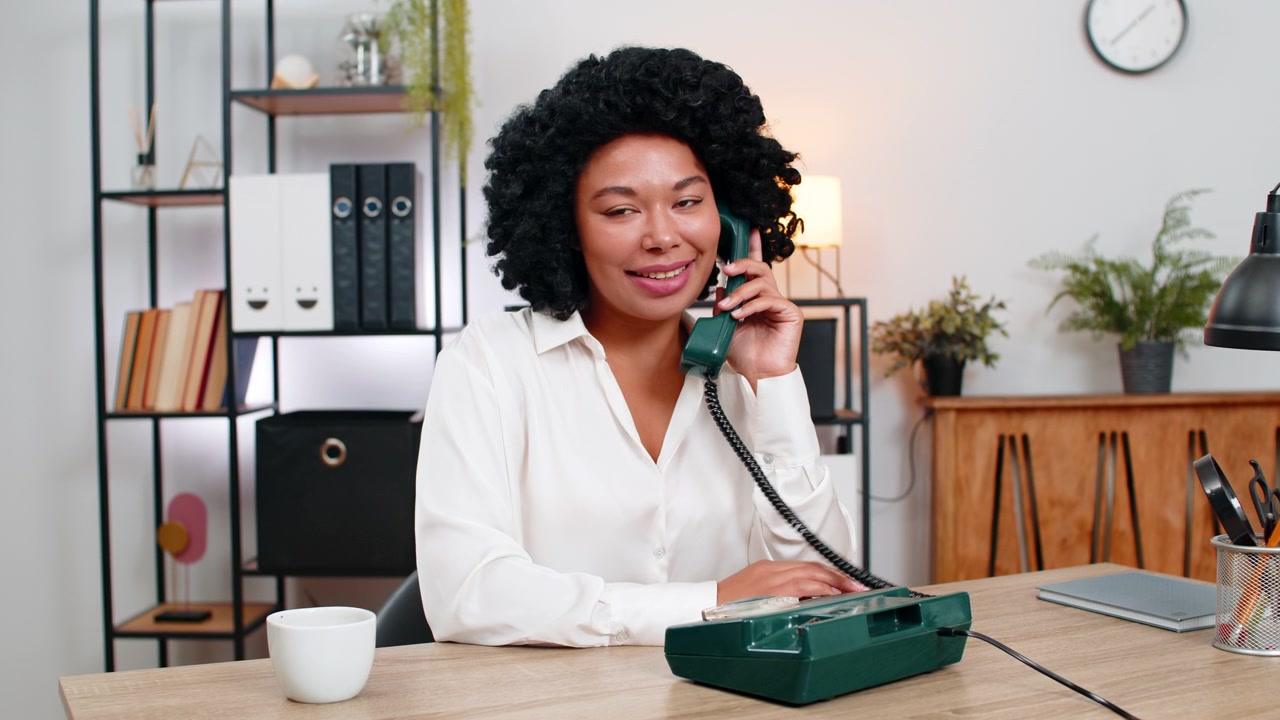 Afroamerikansk forretningskvinne ler mens hun snakker i en retro gammel telefon, og smiler vennlig under en klientsamtale.