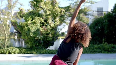 Mujer-Birracial-Feliz-Practicando-Yoga-Estirándose-Junto-A-La-Piscina-En-Un-Jardín-Soleado,-Cámara-Lenta,-Espacio-Para-Copiar