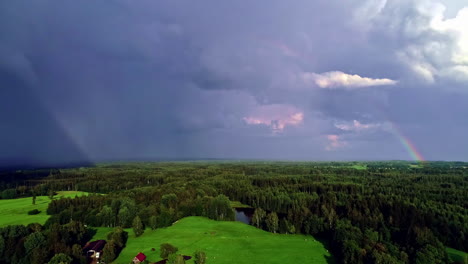 Arco-Iris-En-El-Cielo-Nublado-Oscuro-Sobre-El-Paisaje-Verde-De-árboles-Y-Un-Lago