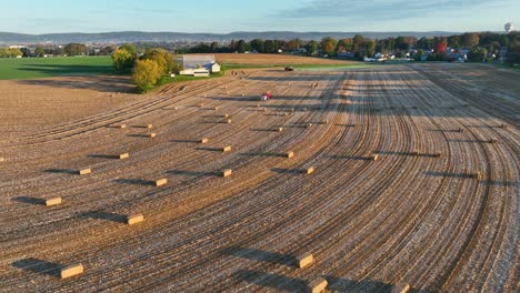 Luftaufnahme-Eines-Ackerlandes-Mit-Gepflegten-Furchen,-Verstreuten-Heuballen-Und-Einem-Traktor-Bei-Sonnenuntergang