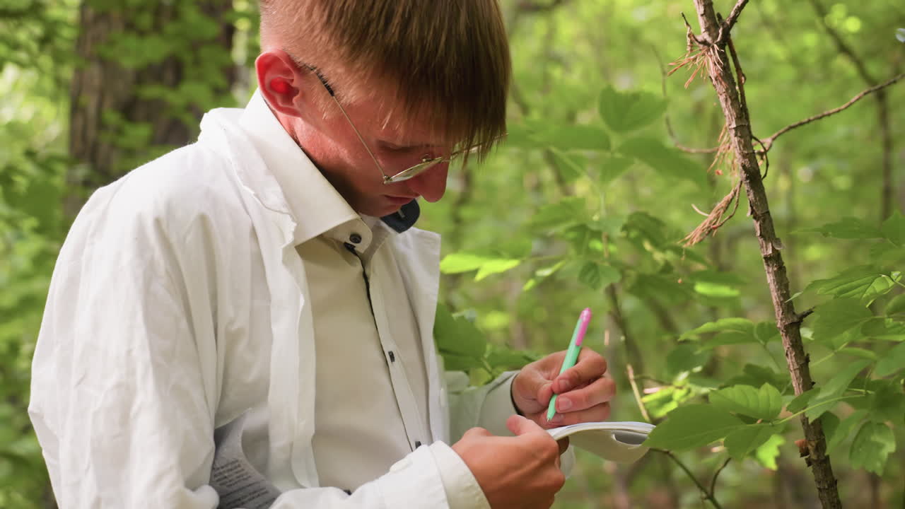 Young botany student in white coat stands in forest under bright sunlight with serious expression writing notes in jotter using pen, documenting natural observations among lush green leaves