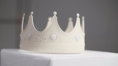 beige crown with ornaments and white pearl jewels with peaks out of plastic falling and landing on a white pedestal