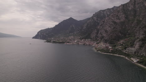 Droneshot-Sobre-La-Ciudad-Limone-Italia-En-El-Lago-De-Garda-En-Un-Día-Gris-Con-Montañas,-Nubes-Y-Troncos-De-Agua