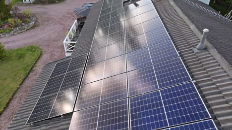 Drone-rising-above-a-house-with-solar-modules,-clouds-mirroring-from-the-surface