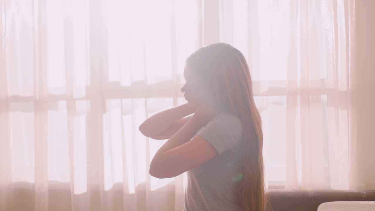Side view of young girl seated near bright window stretching neck gently with both hands due to muscle tension, bathed in soft sunlight, expressing fatigue and calm relief