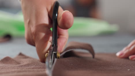 female hand firmly gripping scissors slicing through brown fabric following white chalk guideline on work table, close up view of precision cutting process in tailoring or dressmaking workshop