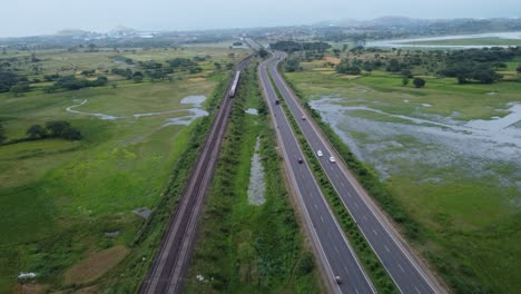 Luftaufnahme-Der-Indischen-Eisenbahnstrecke-Und-Der-Indischen-Autobahn,-Die-In-Einem-Schuss-Parallel-Verlaufen,-Während-Güterzug-Und-Lokomotive-Vorbeifahren