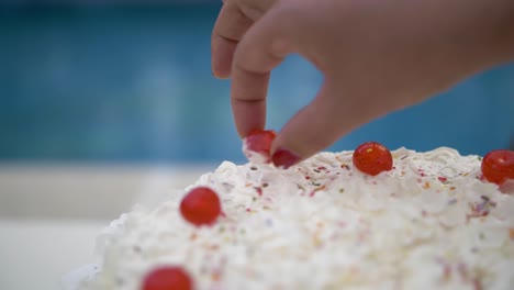 Woman´s-hand-putting-a-red-candied-fruit-in-a-white-cream-cake-dessert-as-decoration,-outdoor-near-the-swimming-pool-in-summer-detail-in-slowmotion