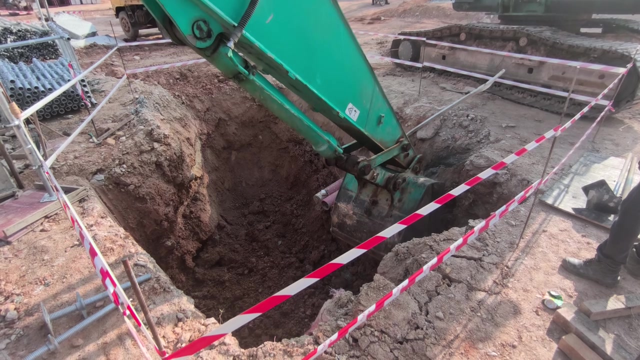 Bir ekskavatör, elektrik menholü kurulumu için derin bir çukur kazıyor. Kazara düşmeleri önlemek amacıyla parlak güvenlik bariyer bandı inşaat alanını çevreliyor.