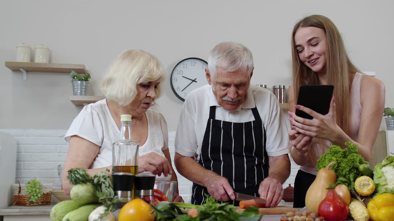una pareja de abuelos mayores cortando verduras para ensalada, escuchando la receta de una chica con una tableta.