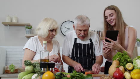 Senior-grandparents-couple-cutting-vegetables-for-salad,-listening-recipe-from-girl-with-tablet