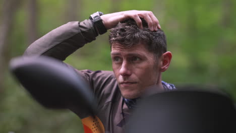 Biker-on-motorcycle-getting-ready-to-ride-in-the-mountains-off-road,-scratching-his-head-and-looking-around