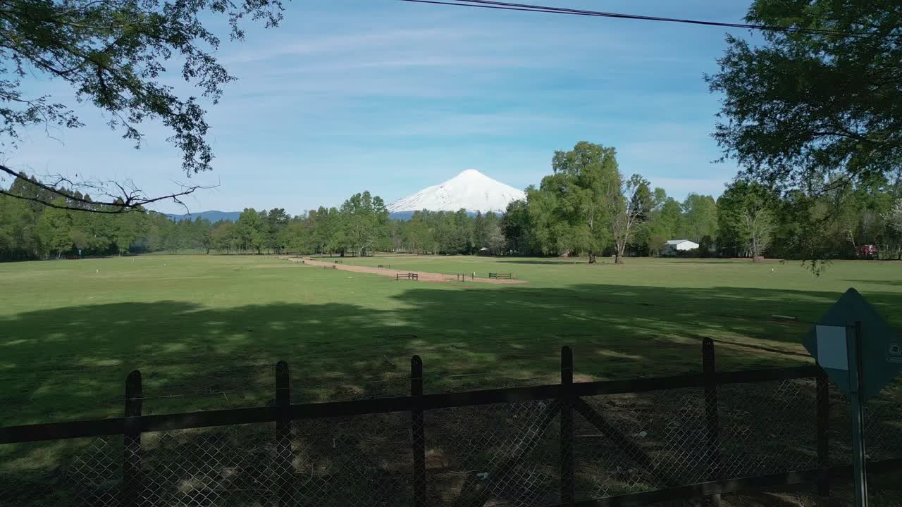 Drone shot of an open green field near Pucón, Chile, with the snow-capped Villarica volcano in the distance framed by trees and clear skies