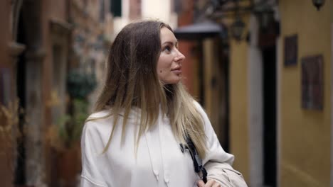 Attractive-woman-walks-along-a-city-narrow-street-with-backpack,-looks-with-interest-around