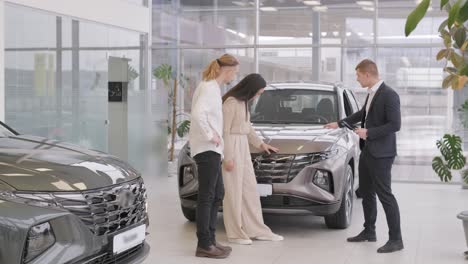 young car salesman showing to young couple new automobile at dealership salon.