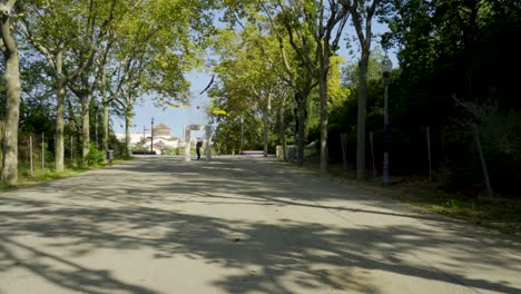 Young-man-with-backpack-standing-and-posing-in-middle-of-park-road-at-Montjuic-National-Palace,-Palau-Nacional-in-Barcelona,-Spain-on-beautiful-sunny-day-with-trees-and-lanterns-in-park-area