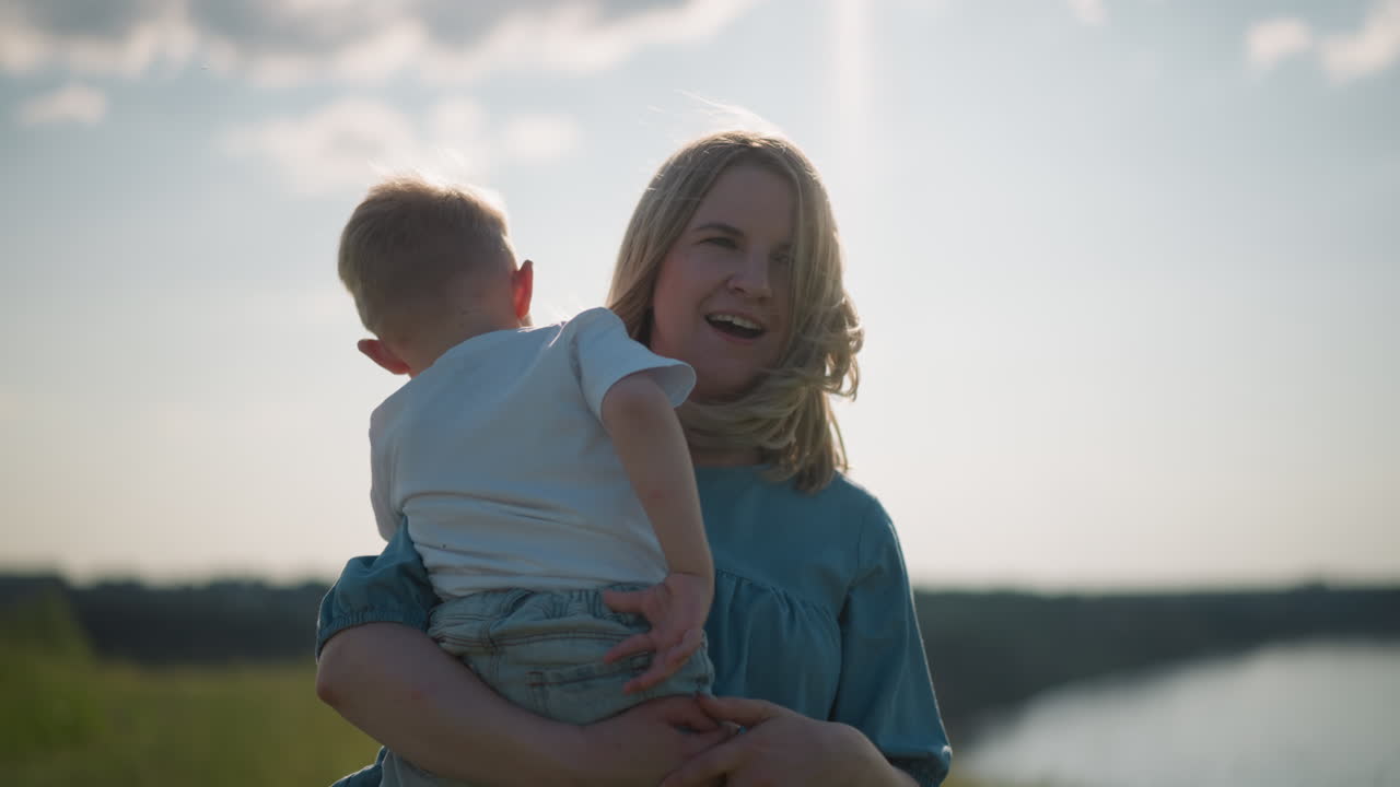una mujer con un vestido azul sostiene a su hijo pequeño, que lleva una camiseta blanca, mientras ambos miran hacia un lago sereno. el momento pacífico captura el vínculo entre madre e hijo