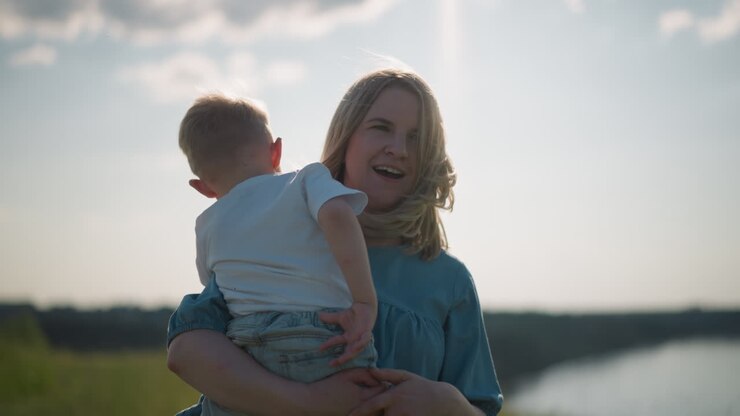 A woman in a blue dress holds her young son, who is wearing a white top, as they both gaze out towards a serene lake. The peaceful moment captures the bond between mother and child