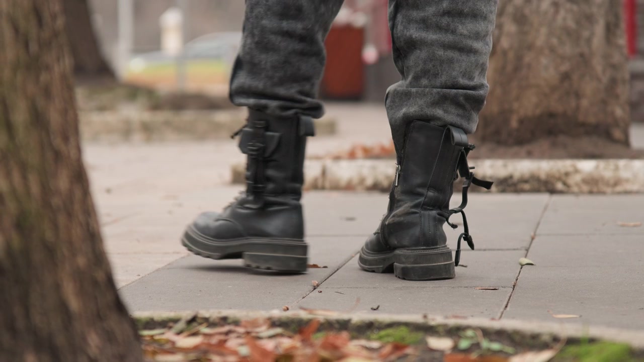 Botas de combate negras caminando por la acera. Primer plano en ángulo bajo de una bota de cuero con cordones pisando el pavimento de hormigón con hojas de otoño esparcidas. El borde de un tronco de árbol en primer plano, bajo una luz nublada y fría.