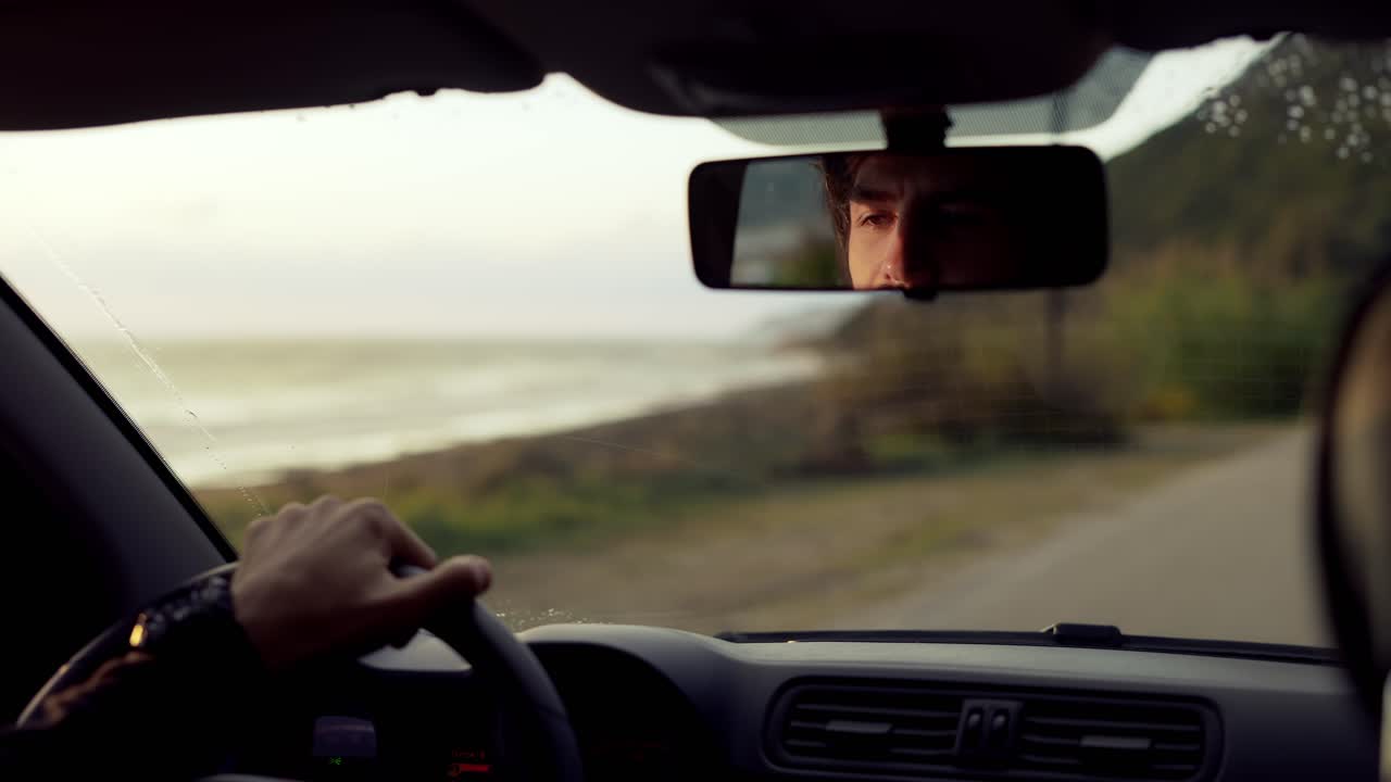 Bearded man driving a car along the coast, focused gaze in rear view mirror, golden hour