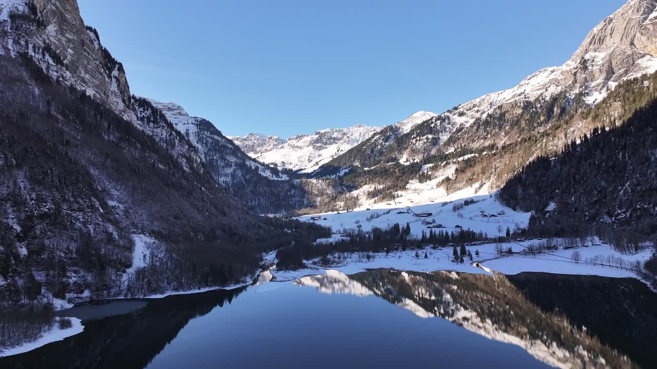 A mesmerizing aerial view of Klöntalersee and the Klöntal valley surrounded by majestic snowy peaks, reflecting on the tranquil lake below. A serene winter landscape in Glarus, Switzerland.