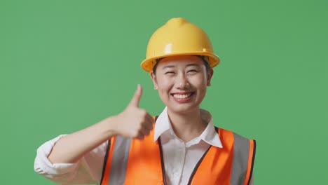 close up of asian female engineer with safety helmet smiling and showing thumbs up gesture to the camera while standing in the green screen background studio