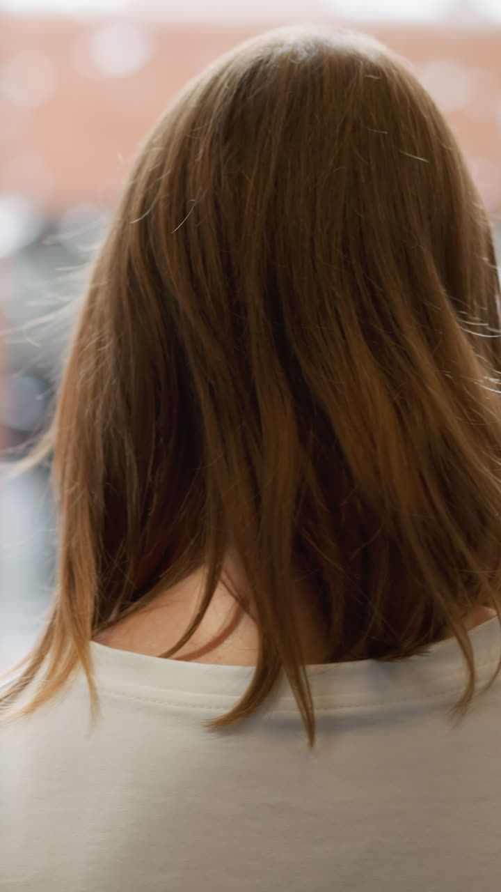 어깨 길이의 갈색 머리카락을 가진 젊은 여성이 색 스웨트 셔츠를 입고, 부드럽게 밝은 배경에서 현대 건축의 흐릿한 패턴과 자연 빛 반사와 함께 고 있습니다.