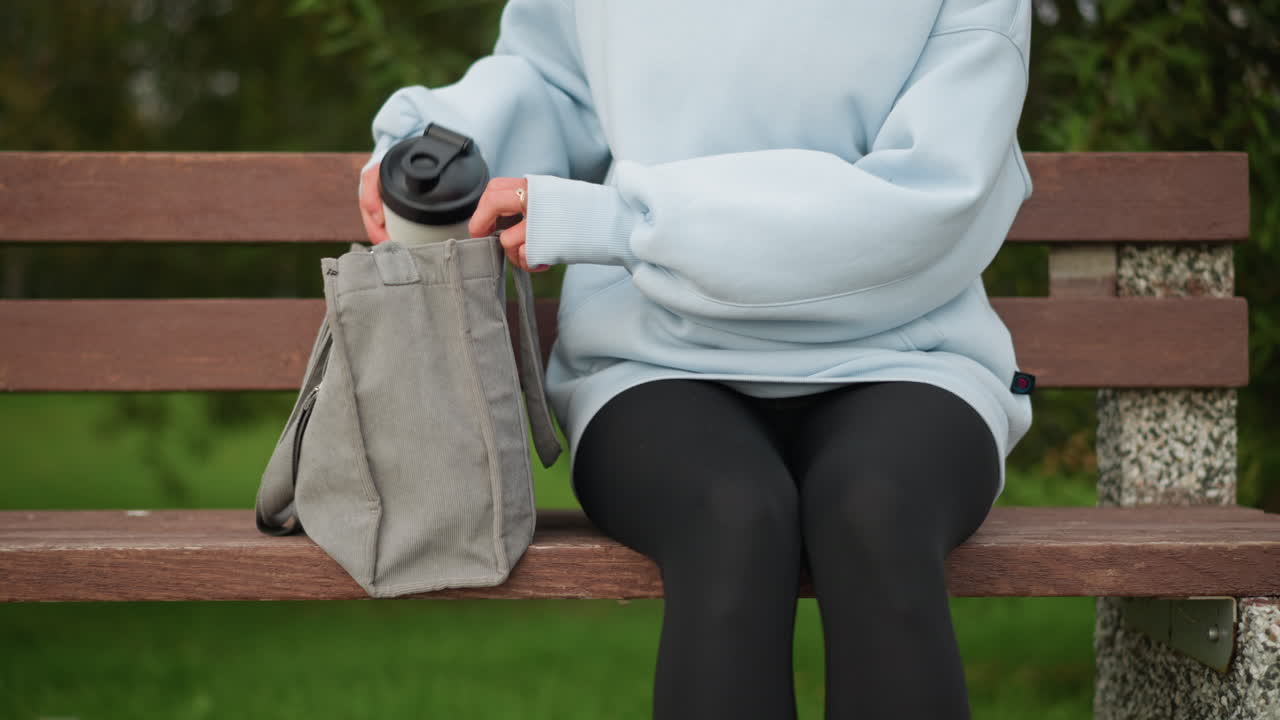 Partial view of girl sitting on bench, pulling water bottle from her bag and opening it in peaceful park setting, ideal for hydration, relaxation, and wellness moments outdoors
