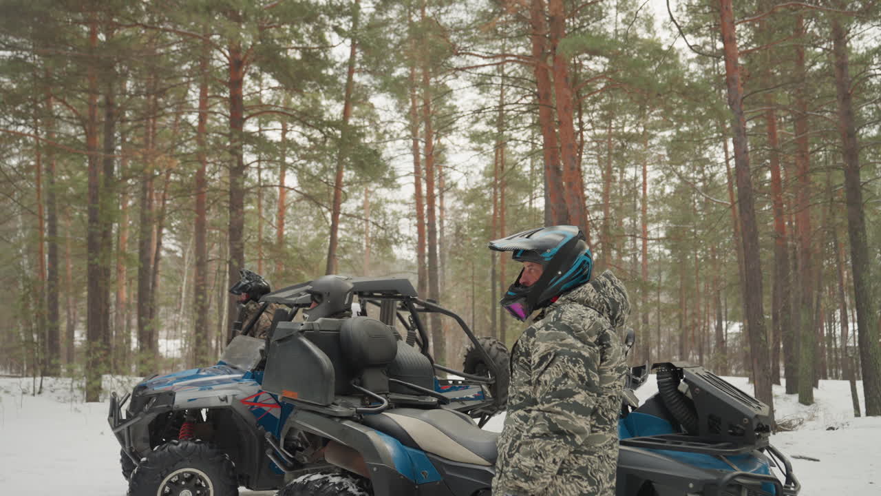 Primo piano di un pilota di quad che regola il cinturino del casco e ispeziona i comandi del quad, tuta mimetica e occhiali, sfondo di pini innevati, postura concentrata, preparazione per un giro fuoristrada estremo, controllo di sicurezza e avviamento del motore.