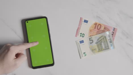 tiro aéreo de notas de 10 e 5 euros ao lado de uma pessoa usando um celular de tela verde