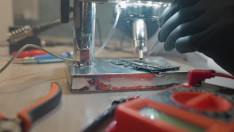 close-up view of a technician using a microscope and precision tools to repair a circuit board, with voltmeter and various equipment on the table