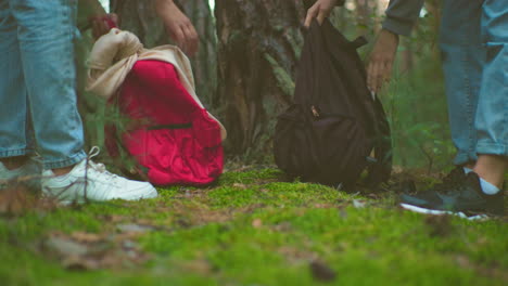 close-up of two women in jeans and sneakers carefully zipping up backpacks retrieved from ground in forest, preparing to continue their hike, with lush greenery and tree trunks in the background