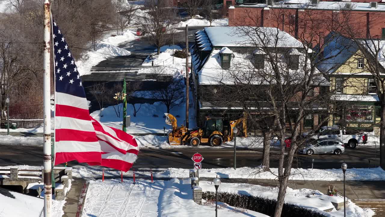 Aerial view of an American town in winter with large U.S. flag waving in bright sunlight. Snow blankets streets and rooftops as vehicles and construction loader move through small city intersection